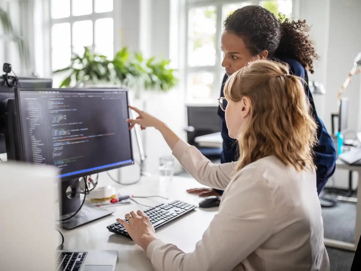 Two developers reviewing code together on a desktop monitor in a bright office.