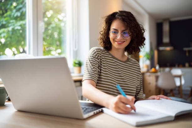 A researcher at a bright desk reviewing sources on a laptop while taking notes in a notebook.